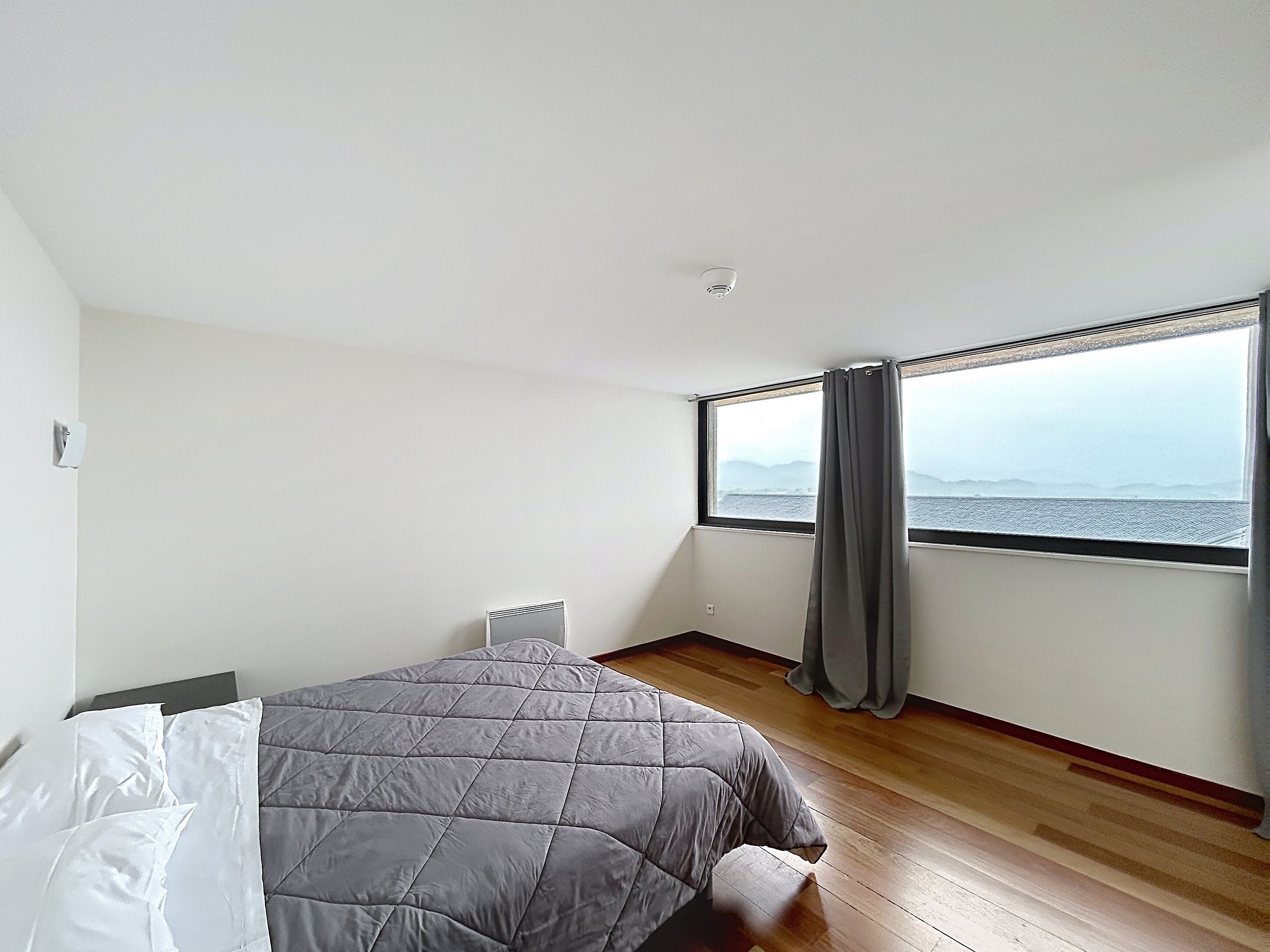 A bedroom with a bed, a window with curtains, a view of the sea, and a white wall.