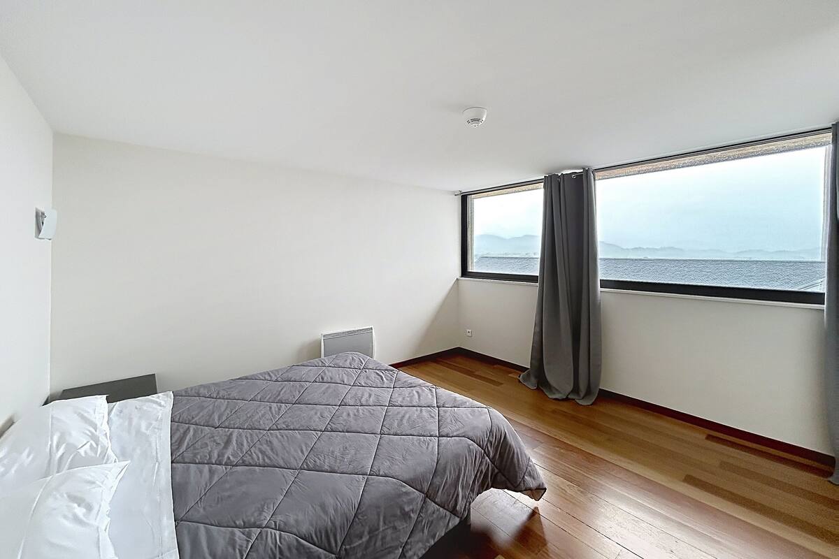 Un dormitorio con cama, ventana con cortinas y vistas al mar, y una pared blanca.