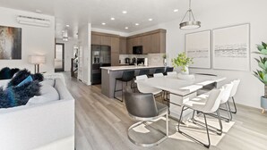 A modern kitchen with a central island, stainless steel appliances, and a dining area with a white table and chairs. - Family Reunion Fun (#13 and #15) SLEEPS 32! GREAT AMENITIES! (St. George)