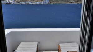 A balcony with two wooden lounge chairs and a small table, overlooking a body of water and a hillside with buildings. - Amorgos the olive garden above the sea I (Katapola)