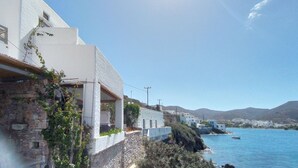 A coastal building with a stone wall and white facade, overlooking a clear blue sea with boats. - Amorgos the olive garden above the sea I (Katapola)