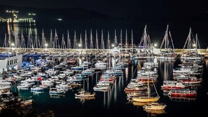 Una marina de noche con numerosos barcos amarrados, un edificio de diseño moderno y una superficie de agua tranquila que refleja las luces.