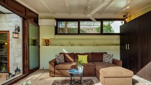 Un espacio de sala acogedor con un sofá seccional marrón, una pequeña mesita de noche con revistas, una planta en maceta y un ventanal con vistas a la vegetación.