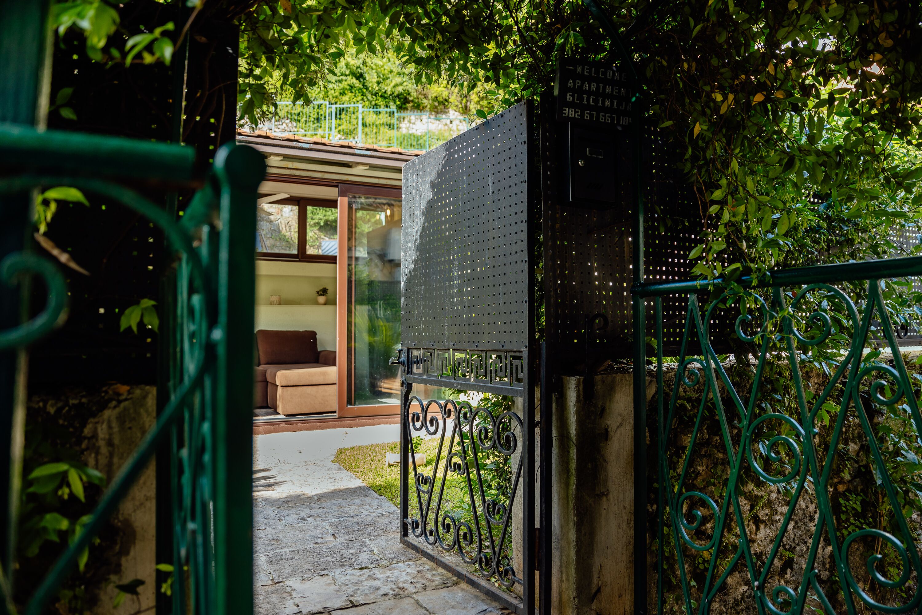 A gated entrance to a residential property with a metal gate, a pathway leading to a glass door, and a small garden area with greenery.