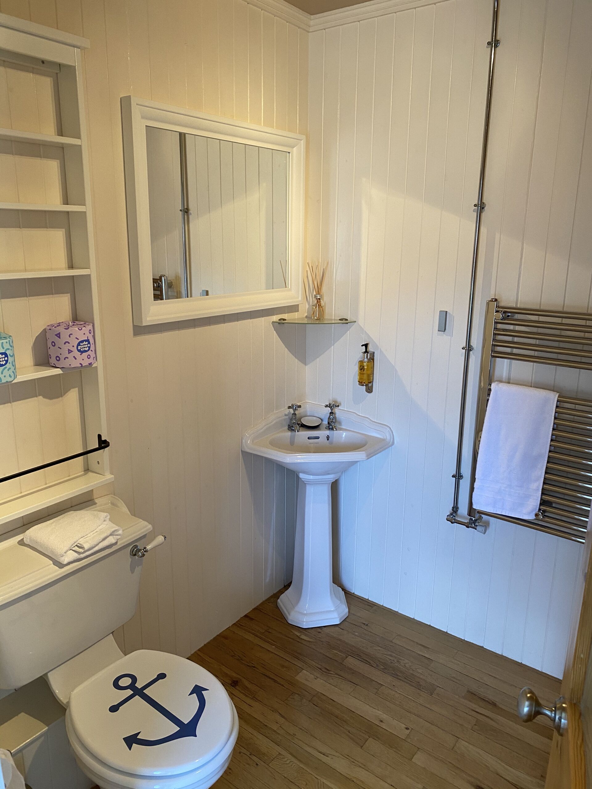 Une salle de bain avec un lavabo blanc, un miroir, des toilettes décorées d’un motif d’ancre, un porte-serviettes et un plancher en bois.