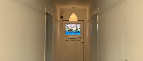 A hallway with a white door at the end, a large white pendant light above, and a small window with a view of the sky and clouds.