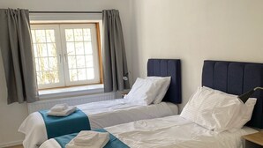 A hotel room with two beds, a window with curtains, a ceiling light, and a small wooden table. - The Crail Maltings (Anstruther)