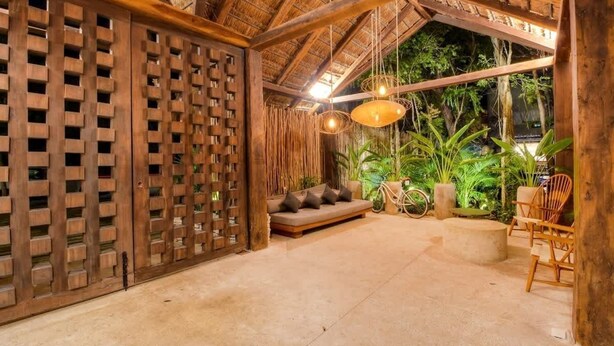 A covered outdoor seating area with a wooden lattice partition, a cushioned bench, hanging light fixtures, and potted plants. - Aldea Zama Condo: Private Pool and Jungle Views (Tulum)