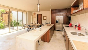 A modern kitchen with a large island, stainless steel appliances, and a sliding glass door leading to a garden. - Aldea Zama Condo: Private Pool and Jungle Views (Tulum)