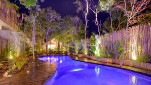 A nighttime swimming pool with a wooden deck, surrounded by tropical plants and bamboo fencing. - Aldea Zama Condo: Private Pool and Jungle Views (Tulum)