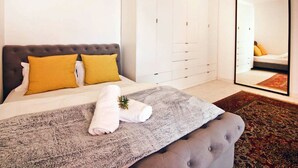Ein Schlafzimmer mit einem Bett, einer grauen Decke, einem grauen, gesteppten Kopfteil, einem weißen Kleiderschrank und einem großen Spiegel.