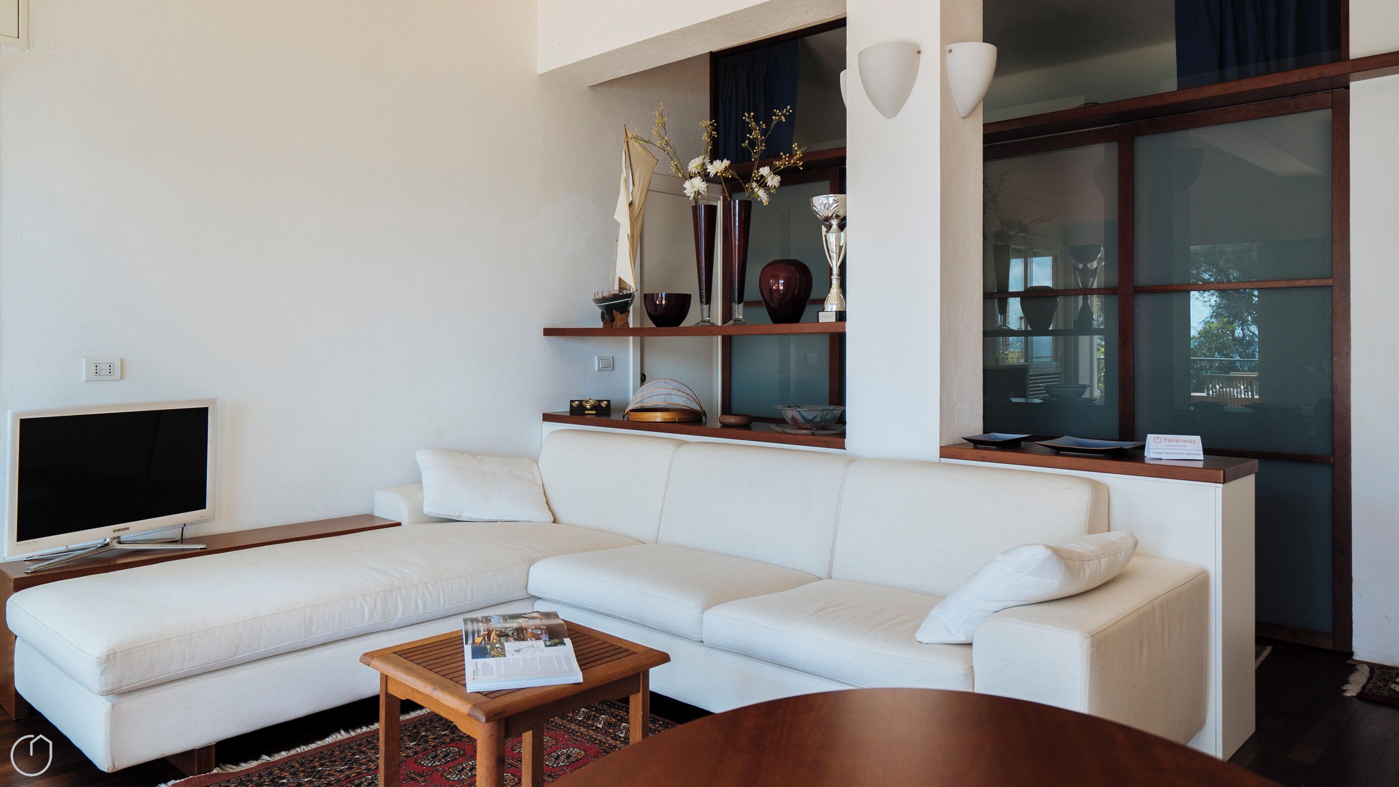 Un salon moderne avec un canapé blanc, une table basse en bois, un téléviseur à écran plat, des objets décoratifs disposés sur des étagères et une grande fenêtre.