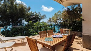 A wooden table with chairs, a book, and a decorative plate on a balcony with a view of trees and a clear sky.