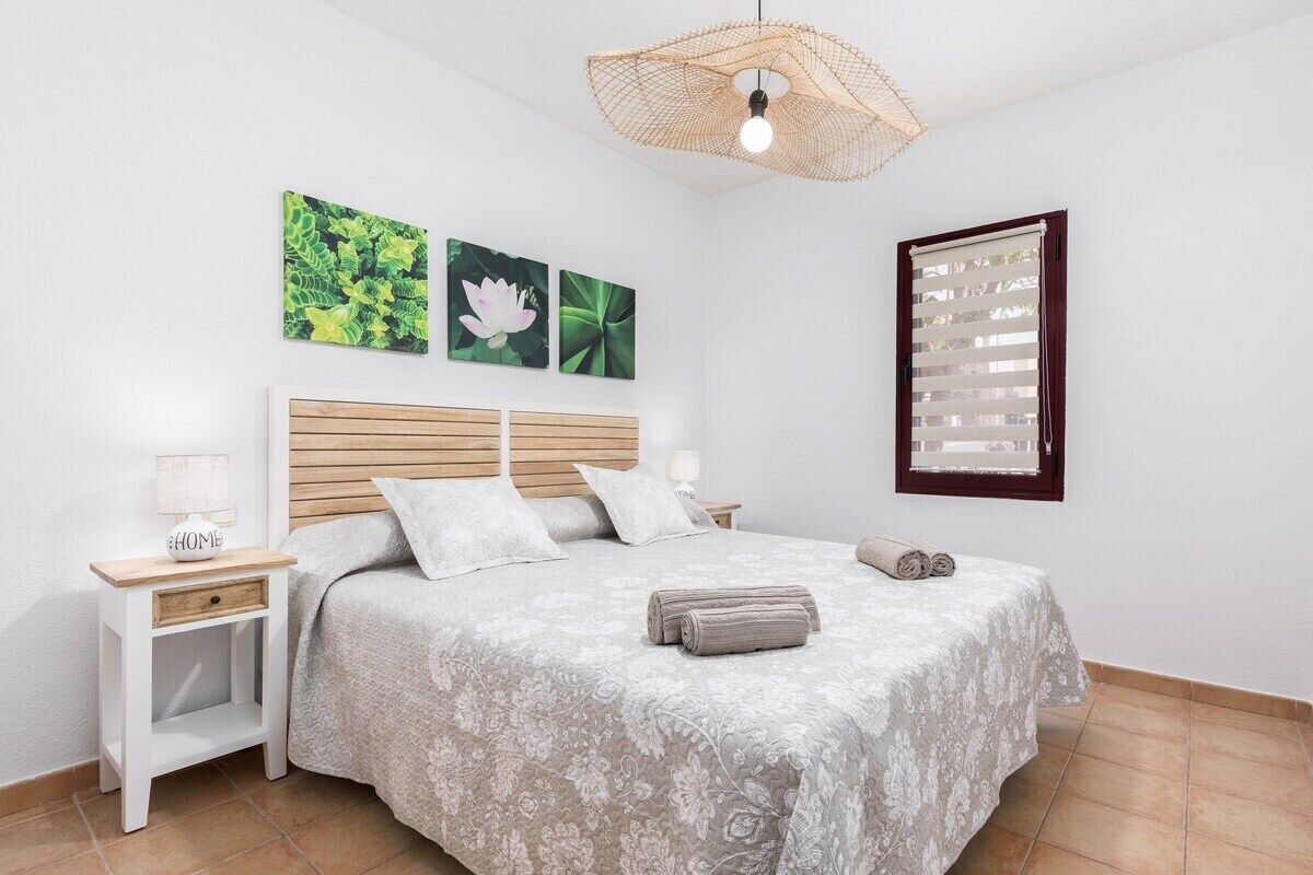A neatly arranged bedroom with a wooden headboard, a bed with a floral bedspread, a small bedside table with a lamp, and a wall-mounted window with blinds.