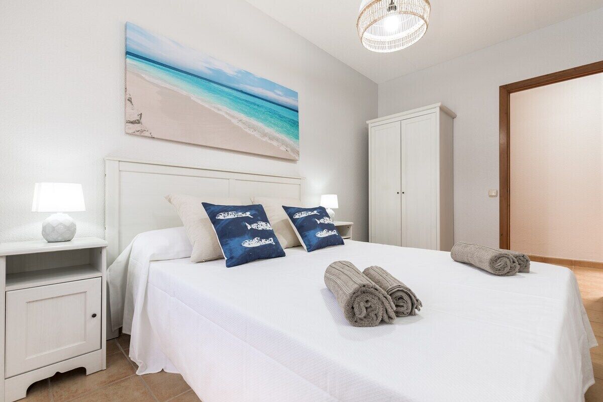 Un dormitorio con una cama, dos mesitas de noche, un armario blanco, un cuadro con motivos playeros y una toalla enrollada sobre la cama.