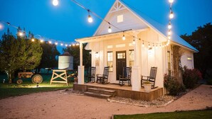 Une maison blanche avec un porche et des guirlandes lumineuses, entourée d’arbres et d’un tracteur.