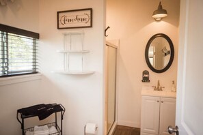 A bathroom with a white vanity, a round mirror, a shelf with a framed sign, a towel rack, and a window with blinds.