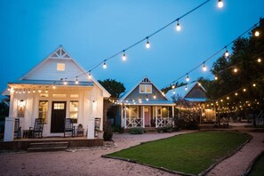 A white building with a porch and string lights, surrounded by a grassy area and trees. - The Gruene Haus at Gruene B&B (New Braunfels)