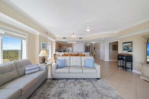 Un salon spacieux avec un canapé, une table basse et un tapis. Au fond, on aperçoit une cuisine équipée de placards et un coin repas.