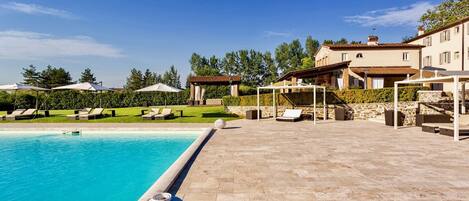 A pool area with lounge chairs and umbrellas, a stone patio, and a building with a tiled roof.
