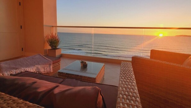 Ein Balkon mit Korbmöbeln, einem kleinen Tisch und Blick aufs Meer im Sonnenuntergang.