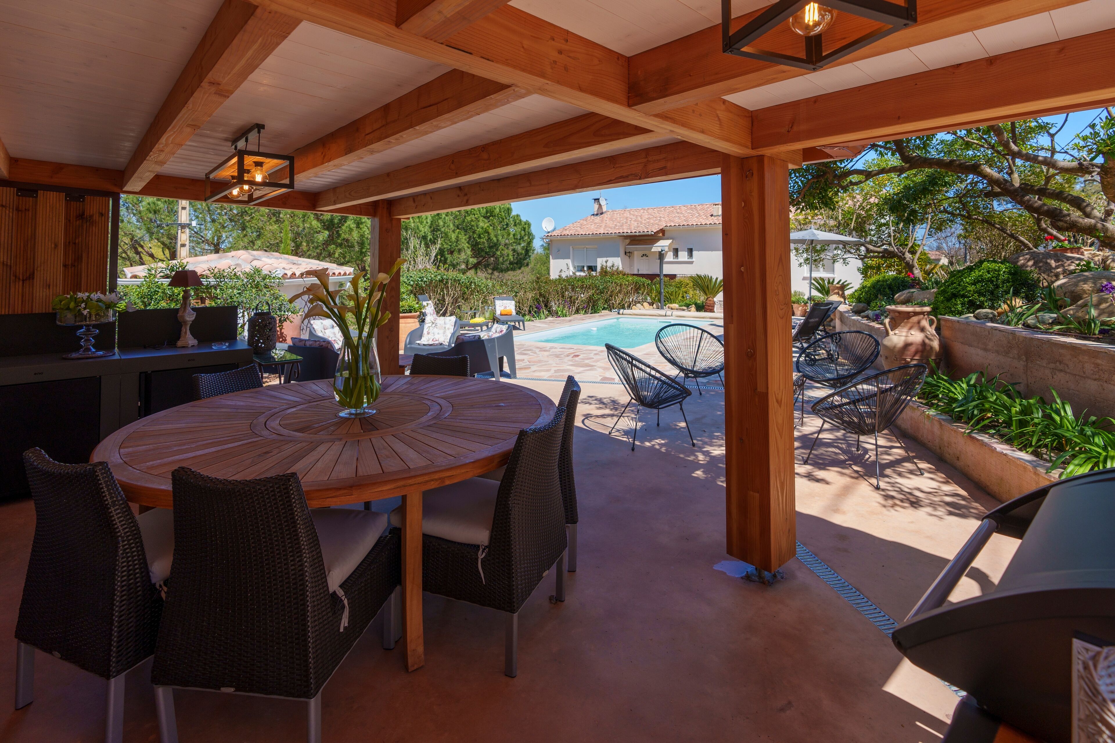 Une terrasse extérieure avec une table ronde en bois et des chaises en osier, une piscine et un mur en pierre.
