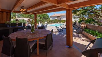 Une terrasse extérieure avec une table ronde en bois et des chaises en osier, une piscine et un mur en pierre.