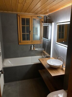 A modern bathroom with a bathtub, a wooden vanity with a sink, and a mirror. - Chalet Soldanella (Saanen)