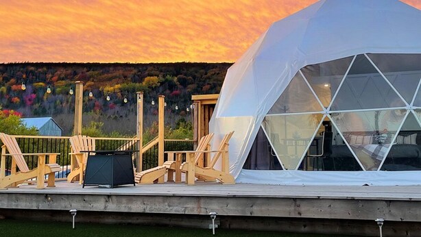 Una estructura de cúpula geodésica con terraza y sillas de madera, con un cielo de atardecer de fondo.