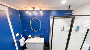 Un baño con pared azul, lavamanos blanco, cabina de ducha de vidrio y espejo hexagonal.