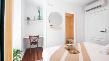 A hotel room with a bed, a desk, a chair, a mirror, a shelf with plants, and a bathroom visible through an open door.