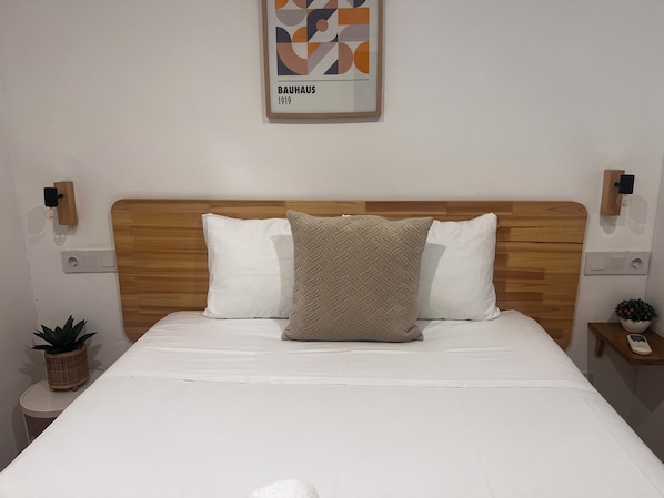 A neatly made bed with a wooden headboard, two white pillows, and a textured beige pillow. There is a framed artwork above the bed, a small plant on a side table, and a bedside table with a lamp and a phone. - Marisol Self Check-in Guesthouse (San Sebastián)