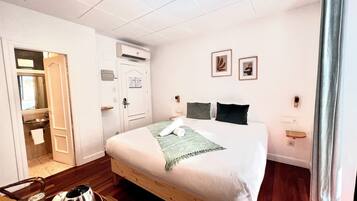 Habitación de hotel con cama, piso de madera, baño con lavamanos y espejo, y una puerta que da a otra habitación.