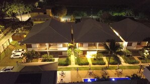 A hotel with multiple buildings, each with a sloped roof and a balcony. There are cars parked in front, and a person walking on the lawn. - Luxury apartment, 3 bedrooms (2 suites) - Praia da Espera - Itacimirim (Camaçari)