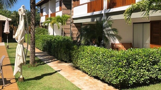 A resort with a palm tree-lined pathway, wooden deck, and a building with a ship's wheel on the wall. - Luxury apartment, 3 bedrooms (2 suites) - Praia da Espera - Itacimirim (Camaçari)