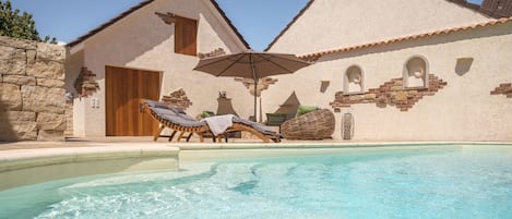 Un espace piscine agrémenté de chaises longues et d’un parasol, entouré d’un bâtiment aux murs en pierre et aux toits recouverts de tuiles.