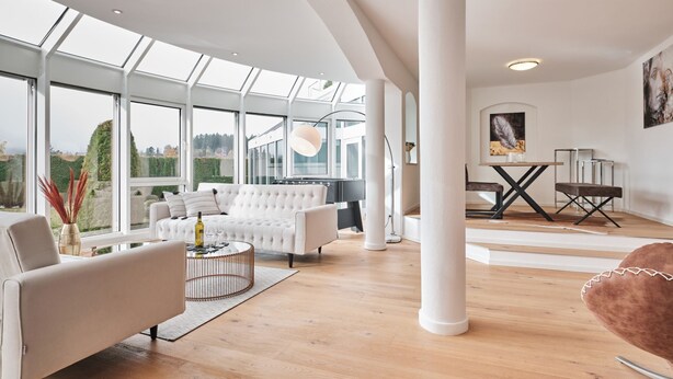 A spacious living area with a white sofa, a coffee table, and a dining table with chairs. Large windows provide natural light and views of the outdoors. - Loft am Schluchsee (Schluchsee)