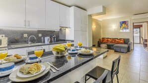 Una cucina moderna con mobili bianchi, piani in granito e un'area pranzo con un divano grigio e un quadro appeso al muro.