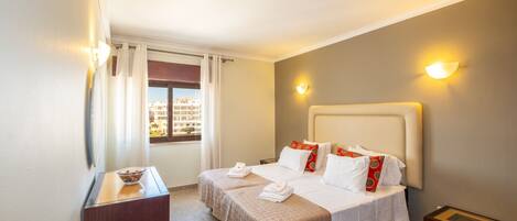 Une chambre d’hôtel avec deux lits, une petite table, une fenêtre avec des rideaux et une vue sur les bâtiments environnants.