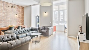 A modern living room with a sectional sofa, a round coffee table, and a TV. There is a brick wall, a large window, and a ceiling light fixture.