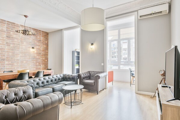 A modern living room with a sectional sofa, a round coffee table, and a TV. There is a brick wall, a large window, and a ceiling light fixture.