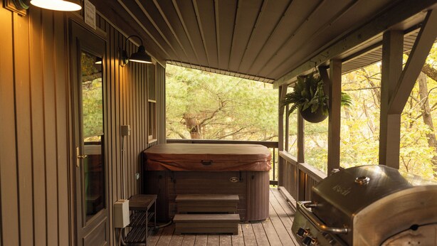 Eine überdachte Veranda mit Whirlpool, Grill und Blick auf Bäume.