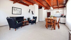 A spacious living area with a wooden ceiling, a black leather sofa, a wooden dining table, and a kitchen in the background. - CAPTAIN'S COTTAGE (Cape Town)