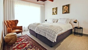 A bedroom with a large bed, a brown leather armchair, a patterned rug, and two wall art pieces. - CAPTAIN'S COTTAGE (Cape Town)