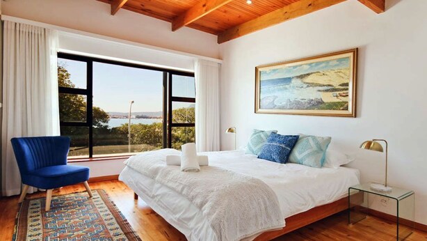 A bedroom with a large bed, a blue chair, a glass side table, and a painting on the wall. - ADMIRAL'S COTTAGE (Cape Town)