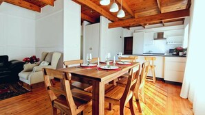 A dining area with a wooden table set for four, surrounded by wooden chairs. A kitchen is visible in the background with white cabinets and a black countertop. - ADMIRAL'S COTTAGE (Cape Town)