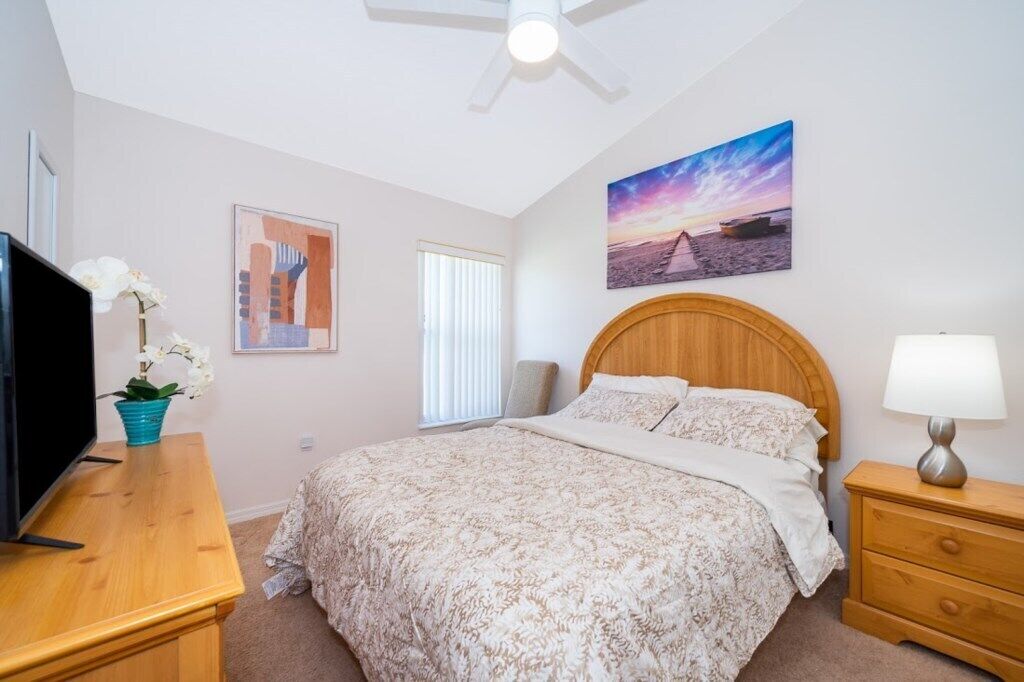 A bedroom with a bed, a TV, a wooden nightstand, a lamp, a ceiling fan, a window with blinds, and a painting on the wall.