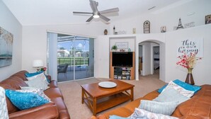 A living room with a brown leather sofa, a wooden coffee table, and a television. There is a sliding glass door leading to a balcony. - Sunset Villa, Minutes from Disney. Exclusive 4 Bed, 2.5 Bath Pool Home (Kissimmee)