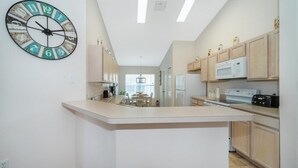 A modern kitchen with a central island, wooden cabinets, a large wall clock, and a microwave. - Sunset Villa, Minutes from Disney. Exclusive 4 Bed, 2.5 Bath Pool Home (Kissimmee)