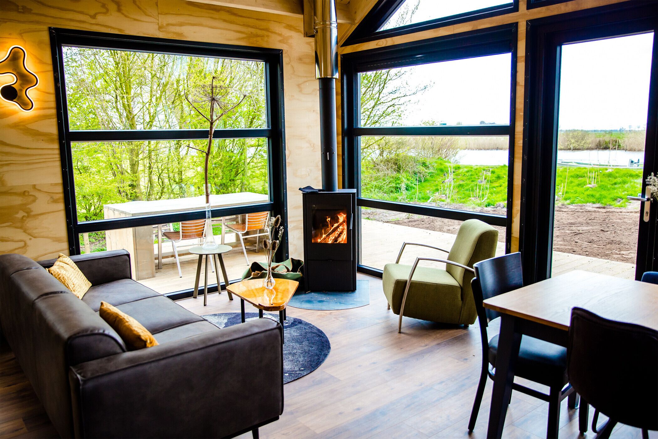 A modern living room with a sofa, chairs, a table, and a wood-burning stove. Large windows offer a view of greenery and a body of water.